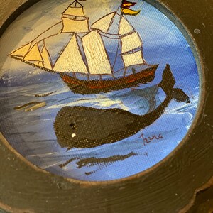K&ouml;nnte beinhalten: Ein gerahmtes Gem&auml;lde mit einem Segelboot mit wei&szlig;en Segeln und einem schwarzen Wal, der darunter schwimmt. Das Gem&auml;lde hat einen blauen Hintergrund und ist unten rechts mit "Hena" signiert.