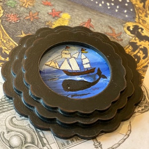 K&ouml;nnte beinhalten: Ein gerahmtes Gem&auml;lde, das ein Schiff zeigt, das auf einem blauen Meer segelt, unter dem eine Wal schwimmt. Das Gem&auml;lde ist vor einem sternenklaren Nachthimmel mit einem gelben Mond platziert.