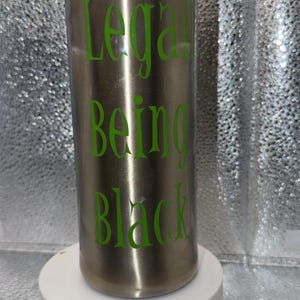 May include: A tall, cylindrical, metallic silver tumbler with a clear plastic lid. The tumbler features bright green text printed vertically on the side that reads "Legally Being Black" in a stylized, serif font. The surface has a smooth finish.