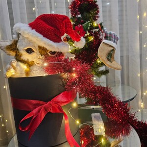 May include: A white and brown crocheted animal wearing a red Santa hat is sitting in a black gift box with a red ribbon. The box is surrounded by red tinsel and a Christmas tree with red ornaments.