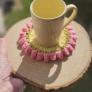 Op de afbeelding: Een gele keramische mok met een handvat staat op een gehaakte onderzetter met roze en groene bloemen. De onderzetter ligt op een houten schijf.