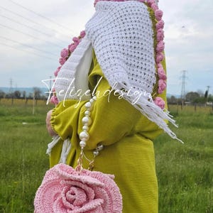 Peut inclure: Un châle blanc en crochet avec des pompons roses et verts sur les bords. Le châle est drapé sur une personne portant une robe jaune et un sac à main en crochet en forme de rose rose.