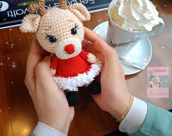 May include: A crocheted reindeer toy wearing a red dress with white trim. The reindeer is being held by a person wearing a green shirt and a gold bracelet. The reindeer is sitting on a wooden table with a cup of coffee and a saucer.