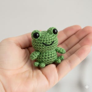 May include: A small, green crocheted frog with large black eyes and a smiling mouth. The handmade amphibian has small arms and legs, and is held in an open hand. The frog is a cute, whimsical toy.