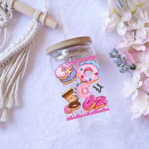 May include: A clear glass tumbler with a bamboo lid and a straw. The tumbler is decorated with colorful donut illustrations and the text "To live like donuts, more donuts". The design includes pink, blue, and brown donuts.
