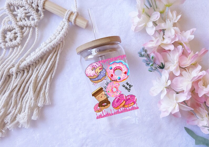 May include: A clear glass tumbler with a bamboo lid and a straw. The tumbler is decorated with colorful donut illustrations and the text "To live like donuts, more donuts". The design includes pink, blue, and brown donuts.