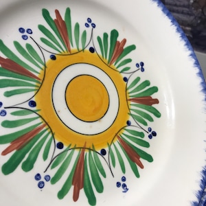 Peut inclure: Assiette en céramique blanche avec un motif peint à la main. L'assiette présente un cercle jaune central avec un anneau intérieur blanc, entouré d'éléments décoratifs verts, marron et bleus. Le bord a une bordure bleue. Cette assiette décorative est idéale pour servir des aliments.