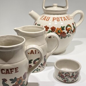 Peut inclure: Un service à thé en céramique crème avec des motifs floraux et de coqs. L'ensemble comprend une théière avec les mots "EAU POTABLE", deux pichets avec "CAFE" et "LAIT", et un petit bol. Les motifs présentent des accents rouges, oranges et verts.