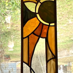 May include: A vertical rectangular stained glass panel featuring a geometric composition of circles and angular shapes in shades of amber, orange, yellow, green, and clear glass, all held together by dark metal lead lines in a grid pattern.