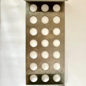 May include: A rectangular metal object with a series of evenly spaced, circular holes. Each hole contains a white, spherical object. The metal frame has a brushed finish and a handle at each end. The object is likely a kitchen or household tool.