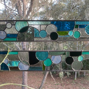 May include: Two stained glass panels featuring geometric shapes in shades of blue, green, and black. The panels are suspended from chains and framed in metal.