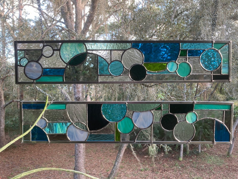 May include: Two stained glass panels featuring geometric shapes in shades of blue, green, and black. The panels are suspended from chains and framed in metal.