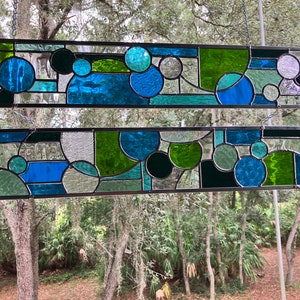 May include: Two stained glass panels featuring geometric shapes in shades of blue, green, and black. The panels are suspended from chains and framed in black metal.