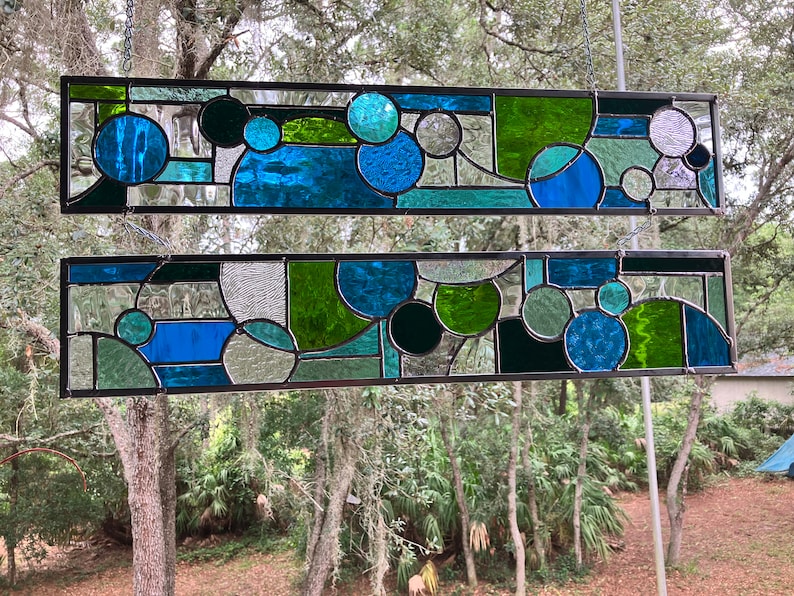 May include: Two stained glass panels featuring geometric shapes in shades of blue, green, and black. The panels are suspended from chains and framed in black metal.