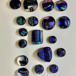 May include: An assortment of dichroic glass cabochons in various shapes, including round, square, and teardrop. The glass pieces feature iridescent colors of blue, purple, and teal, with some having pink and green accents. The cabochons are arranged on a white surface.