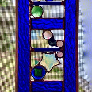 May include: A stained glass window hanging with a blue background and a copper frame. The window features a design of stars and circles in various colors, including green, pink, and white.
