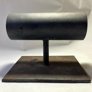 May include: A dark gray metal T-shaped display stand featuring a horizontal cylindrical bar supported by a vertical post attached to a flat square base. The metal surface shows signs of wear and oxidation with visible dark spots and texture.