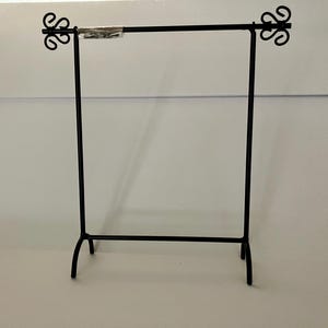 May include: A black metal display rack featuring a horizontal bar supported by two vertical posts with curved feet. The top ends of the horizontal bar are finished with decorative scrollwork accents, and a small plastic bag is attached to the bar.