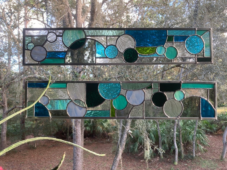 May include: Two stained glass panels with a geometric design featuring circles and squares in shades of blue, green, and grey.