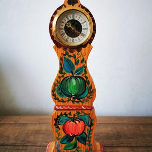 May include: A wooden clock with a painted design of apples and leaves. The clock face has Roman numerals and is surrounded by a gold-coloured trim.