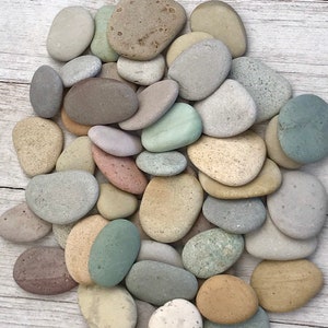 May include: A collection of smooth, rounded stones in various shades of gray, beige, and green. The stones are arranged in a pile on a white wooden surface.