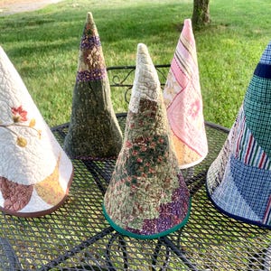 May include: Five cone-shaped fabric covers with quilted textures. One is white with floral embroidery, one is dark green with floral patterns, one is a green and purple patchwork, one is pink and white striped, and one is a blue and white plaid patchwork.