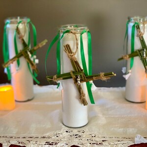 May include: Three white candles with green and brown ribbon decorations. Each candle has a woven cross made of green twigs and brown twine.