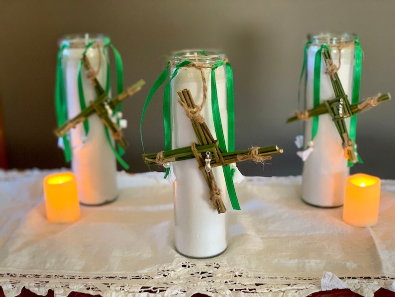 May include: Three white candles with green and brown ribbon decorations. Each candle has a woven cross made of green twigs and brown twine.
