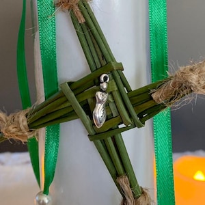 May include: A green woven cross with a silver charm in the center. The charm is a stylized female figure with a spiral design.
