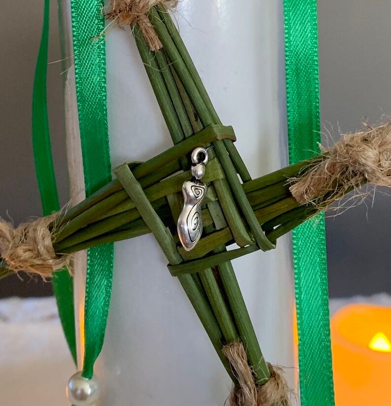 May include: A green woven cross with a silver charm in the center. The charm is a stylized female figure with a spiral design.
