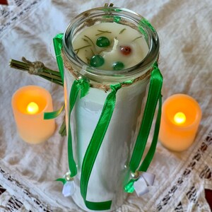 May include: A white candle with green ribbon and green and red accents in a clear glass jar. The candle is surrounded by two small, lit candles.