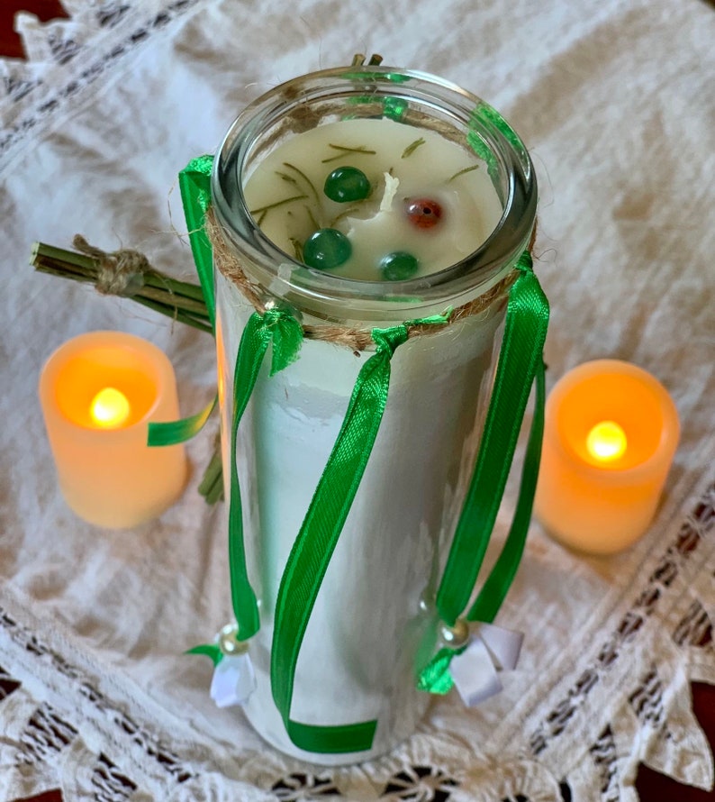 May include: A white candle with green ribbon and green and red accents in a clear glass jar. The candle is surrounded by two small, lit candles.
