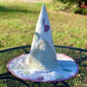 May include: A quilted fabric hat with a tall, pointed crown and a wide, circular brim. The hat features a patchwork design in shades of white, light green, and purple, with a solid pink fabric trim along the edge of the brim.
