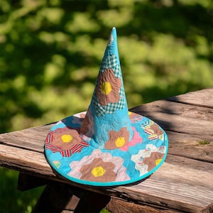 May include: A conical hat constructed from quilted fabric featuring a bright turquoise base color. The surface is decorated with a patchwork pattern of floral shapes in shades of brown, pink, and white, with small yellow centers in each flower.