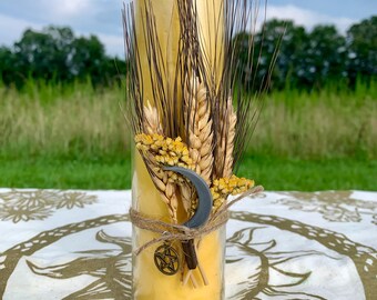May include: A tall, yellow candle in a clear glass jar, decorated with wheat stalks, dried yellow flowers, and a crescent moon charm. The candle is tied with twine and has a pentagram charm. The background is a field and sky.