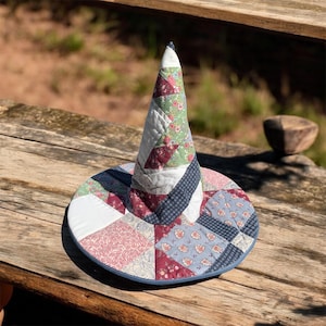 May include: A conical hat constructed from quilted fabric patches in various floral and geometric patterns. The patchwork features shades of sage green, dusty rose, navy blue, and white, with a wide circular brim and a pointed top section.