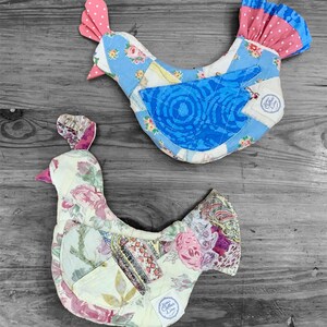 May include: Two stuffed fabric chicken ornaments featuring patchwork designs. The top chicken is blue with floral patterns and a pink polka-dot comb. The bottom chicken is cream with floral and paisley prints. Both have a small white circular label.