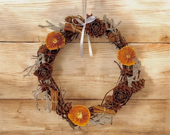 May include: A circular wreath made of twigs, pinecones, dried orange slices, cinnamon sticks, and star anise. The wreath is decorated with a white ribbon for hanging. The background is a light brown wooden surface.
