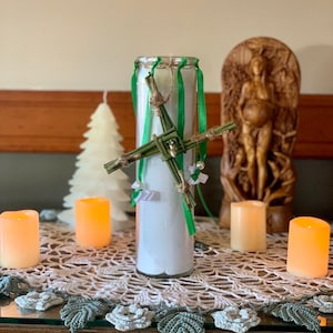 May include: A white candle with a green and brown woven cross decoration, surrounded by four small candles on a white crocheted doily.