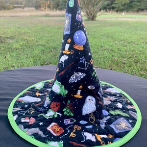 May include: A black quilted witch hat with a wide brim and a pointed top. The hat is decorated with various mystical symbols, including a crystal ball, candles, and stars. The brim is trimmed with a bright green border.