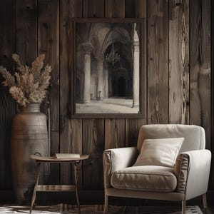 The watercolor architectural scene is displayed in a dark wooden frame with the moody church art. There is an armchair, books and tree stump the room on display. The painting adds a vintage energy to the room.