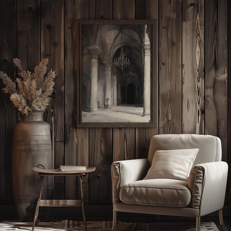 The watercolor architectural scene is displayed in a dark wooden frame with the moody church art. There is an armchair, books and tree stump the room on display. The painting adds a vintage energy to the room.