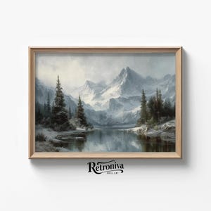 May include: A framed landscape painting depicts a serene winter scene. Snow-covered mountains rise above a calm lake, with evergreen trees lining the shores. The artwork is in a wooden frame, and the overall color palette is muted with shades of gray and white.