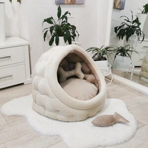 May include: A white and beige dog bed shaped like a dome with a plush, quilted exterior. The bed has a removable cushion and a bone-shaped toy inside. The bed is sitting on a white fluffy rug.