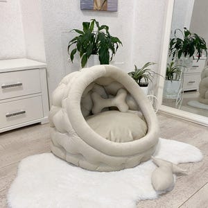May include: A beige dog bed shaped like a cave with a bone-shaped pillow inside. The bed is on a white fluffy rug.