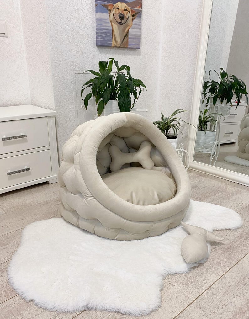 May include: A beige dog bed shaped like a cave with a bone-shaped pillow inside. The bed is on a white fluffy rug.