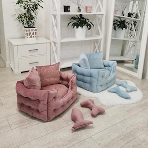 May include: Two plush pet beds, one pink and one blue, with matching bone-shaped toys. The pink bed has a pillow embroidered with "Of True Diamonds". The beds are on a light wood floor, with a white rug and shelves in the background.