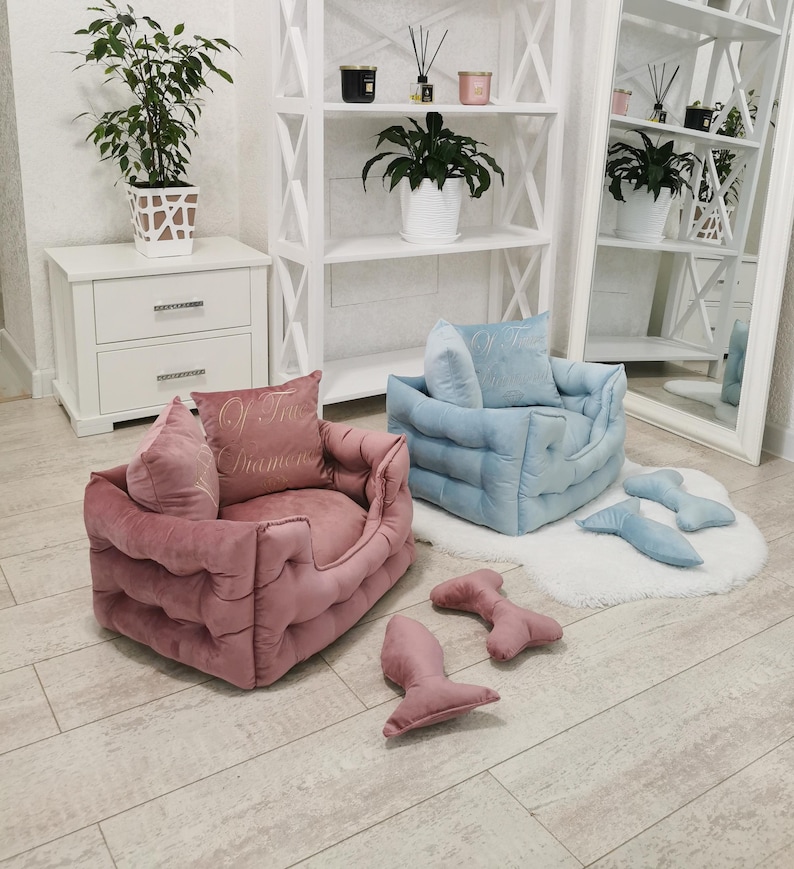 May include: Two plush pet beds, one pink and one blue, with matching bone-shaped toys. The pink bed has a pillow embroidered with "Of True Diamonds". The beds are on a light wood floor, with a white rug and shelves in the background.