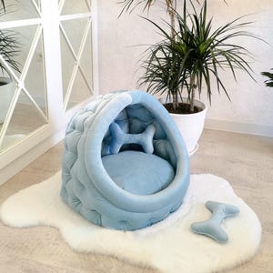 May include: A light blue dog bed shaped like a cave with a plush bone pillow inside. The bed is on a white fluffy rug with a matching bone-shaped pillow.