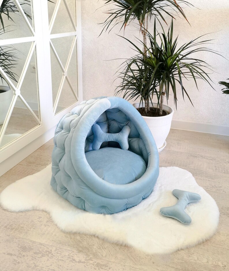 May include: A light blue dog bed shaped like a cave with a plush bone pillow inside. The bed is on a white fluffy rug with a matching bone-shaped pillow.